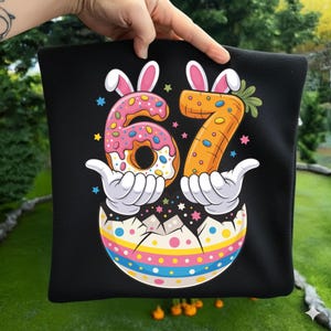 Pode incluir: Fronha preta com um gráfico colorido com tema de Páscoa. O design apresenta o número 67, com o 6 em forma de donut e o 7 como uma cenoura, ambos com orelhas de coelho. Os números são segurados por mãos de desenho animado saindo de um ovo rachado.