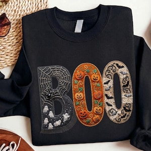Peut inclure: Un sweat-shirt noir à col rond avec le mot BOO imprimé en grandes lettres. Le B est gris foncé avec une toile d'araignée, une araignée et des fantômes blancs. Le O est orange avec des citrouilles et des vignes. Le dernier O est beige avec des chauves-souris noires et des motifs en dentelle.