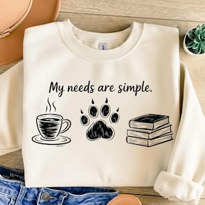 May include: Cream-colored sweatshirt with the phrase "My needs are simple." printed in black. The design includes a coffee cup, a paw print, and a stack of books, all in black. The sweatshirt is folded.
