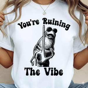 Może przedstawiać: Biała koszulka z czarną grafiką żaby w okularach przeciwsłonecznych i pokazującej środkowy palec. Tekst "You're Ruining The Vibe" znajduje się nad i pod ilustracją żaby.
