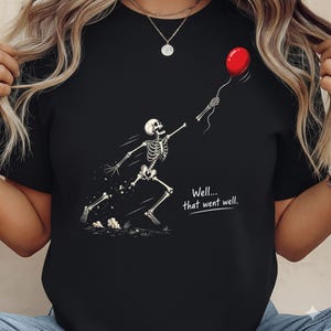 May include: A black t-shirt featuring a graphic of a white skeleton running and reaching for a floating red balloon. The skeleton appears to be losing a hand as it reaches. The text "Well... that went well." is printed in white below the skeleton.