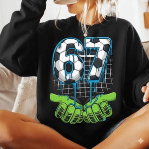 May include: Black sweatshirt with a soccer-themed graphic. The graphic features the number "67" in a soccer ball design with blue dripping accents, held by green goalie gloves. A white grid pattern is in the background.