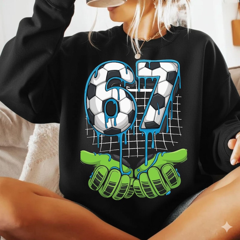 May include: Black sweatshirt with a soccer-themed graphic. The graphic features the number "67" in a soccer ball design with blue dripping accents, held by green goalie gloves. A white grid pattern is in the background.