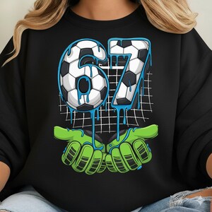 May include: Black sweatshirt with a soccer-themed graphic. The number "67" is designed with a soccer ball pattern and blue dripping accents. Below, green goalie gloves appear to be catching the number. A grid pattern is in the background.