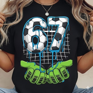 May include: Black t-shirt featuring a soccer-themed graphic. The design includes the number "67" in a soccer ball pattern with blue dripping accents. Below the numbers are green goalie gloves. A white grid pattern is in the background.