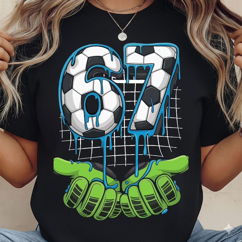 May include: Black t-shirt featuring a soccer-themed graphic. The design includes the number "67" in a soccer ball pattern with blue dripping accents. Below the numbers are green goalie gloves. A white grid pattern is in the background.