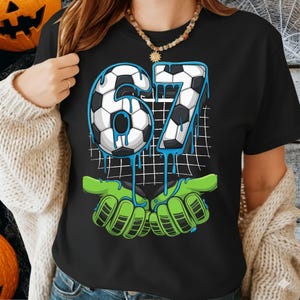 May include: Black t-shirt featuring the number "67" in a soccer ball design with blue dripping accents. The number is set against a white grid, with green hands at the bottom.