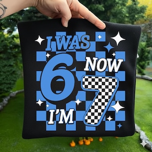 Puede incluir: Cojín cuadrado negro con el texto "I WAS 6 NOW I'M 7" en blanco y azul. El número 6 es azul, y el número 7 es un patrón de tablero de ajedrez en blanco y negro. El fondo es un tablero de ajedrez azul y negro.