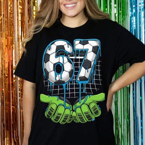 May include: Black t-shirt featuring a soccer ball design with the number 67 in a soccer ball pattern, with blue dripping accents. The design includes green goalie gloves. The shirt is a casual style.