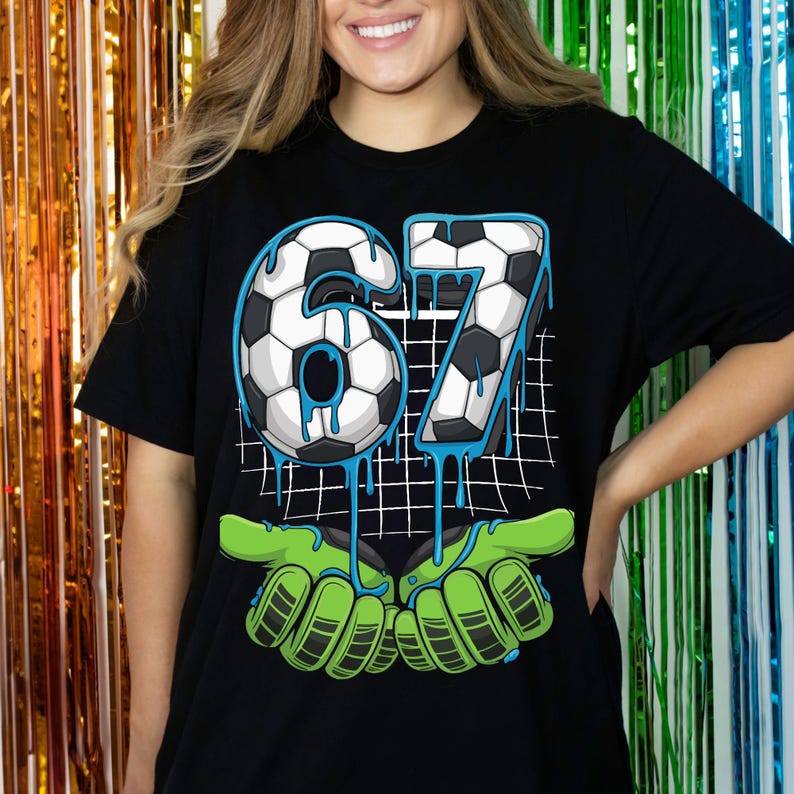 May include: Black t-shirt featuring a soccer ball design with the number 67 in a soccer ball pattern, with blue dripping accents. The design includes green goalie gloves. The shirt is a casual style.