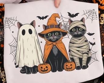 Op de afbeelding: Een wit t-shirt met een afbeelding van drie zwarte katten verkleed voor Halloween. Eén kat draagt een wit spooklaken, één een oranje heksenhoed en cape, en één is gewikkeld in mummiezwachtels. Twee kleine oranje pompoenen, vleermuizen en spinnenwebben omringen hen.