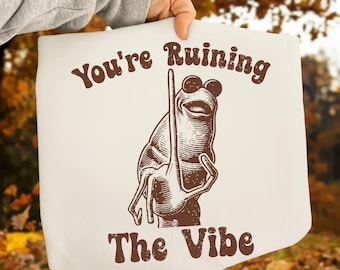 May include: Off-white tote bag featuring a brown illustration of a frog holding a long object. The text "You're Ruining The Vibe" is printed in brown above and below the frog illustration.