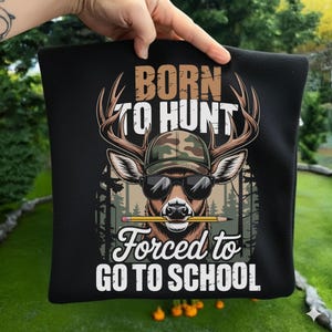May include: Black square pillow with a graphic of a deer wearing sunglasses and a camouflage hat, holding a pencil in its mouth. The text reads "BORN TO HUNT" above and "FORCED TO GO TO SCHOOL" below.