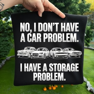Può includere: Cuscino nero con la scritta "NO, I DON'T HAVE A CAR PROBLEM. I HAVE A STORAGE PROBLEM." Sotto il testo ci sono quattro illustrazioni di auto. Il cuscino è tenuto da una mano.