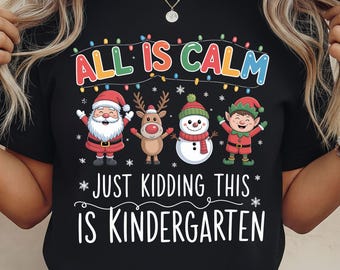 May include: Black t-shirt with a colorful graphic featuring Santa Claus, a reindeer, a snowman, and an elf. The text reads "ALL IS CALM" above the characters and "JUST KIDDING THIS IS KINDERGARTEN" below.