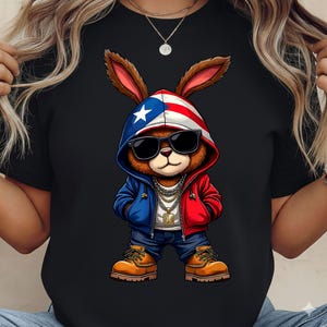 May include: Black t-shirt featuring a cartoon rabbit wearing sunglasses, a red, white, and blue hoodie, blue jeans, and tan work boots. The rabbit has a gold chain and pendant. The hoodie has a star on the hood.