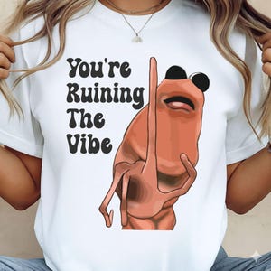 Może przedstawiać: Biała koszulka z czarnym napisem "You're Ruining The Vibe" i kreskówkową ilustracją stworzenia podobnego do krewetki w czarnych okularach. Postać pokazuje środkowy palec.
