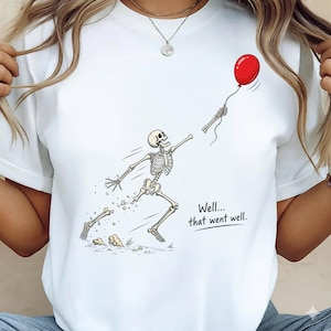 May include: A white t-shirt featuring a graphic of a skeleton running while reaching for a floating red balloon. The skeleton appears to be losing a hand, which is attached to the balloon string. The text reads, Well... that went well.