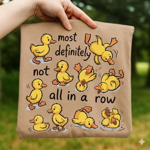 Op de afbeelding: Een bruine draagtas met cartoon gele eendjes in verschillende poses. De tekst op de tas luidt "most definitely not all in a row". De eendjes staan in verschillende posities, sommige zwemmen, sommige staan en sommige zijn ondersteboven.