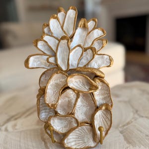 May include: A decorative pineapple sculpture made of white and gold-painted seashells. The pineapple has a textured surface and two small gold hooks. The top of the pineapple is white with gold trim.