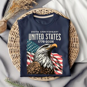 May include: Navy blue t-shirt featuring a detailed illustration of an American bald eagle with the US flag in the background. The shirt commemorates the 250th anniversary of the United States, with the years 1776-2026 printed above the eagle.