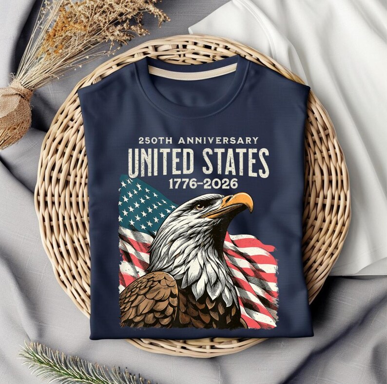 May include: Navy blue t-shirt featuring a detailed illustration of an American bald eagle with the US flag in the background. The shirt commemorates the 250th anniversary of the United States, with the years 1776-2026 printed above the eagle.
