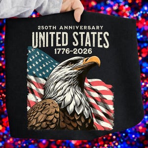 May include: Black apron featuring a detailed illustration of an American bald eagle with the US flag in the background. The text "250th Anniversary United States 1776-2026" is printed at the top. The apron is held up by a person.