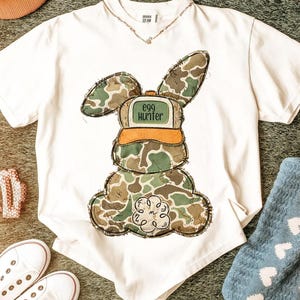 May include: White t-shirt featuring a camouflage bunny graphic with the text "Egg Hunter" on the hat. The bunny design is in shades of green, brown, and tan. The shirt is on a green surface with white sneakers and a blue towel.