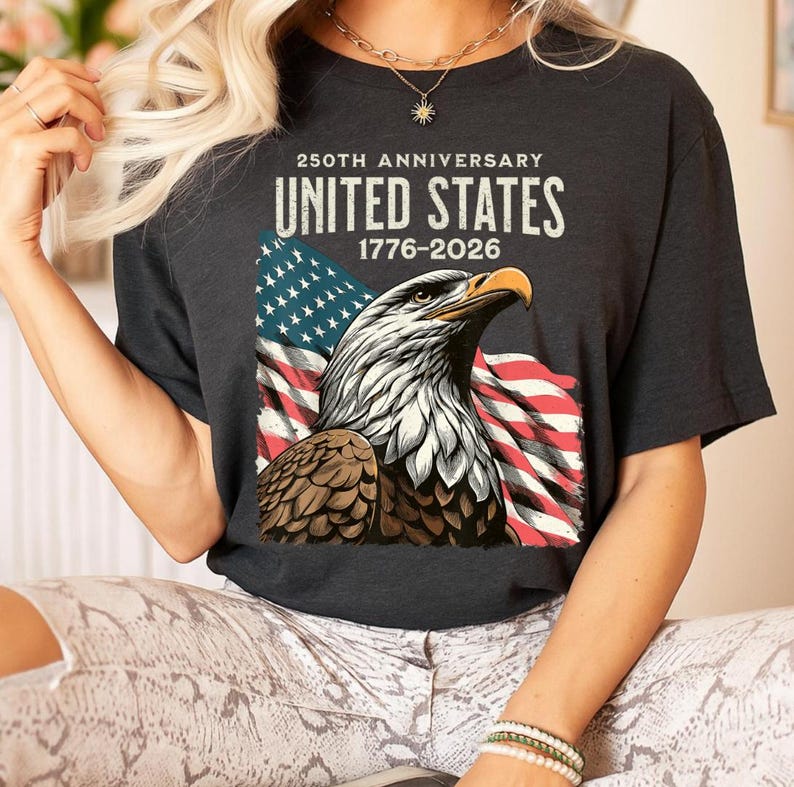 May include: Dark gray t-shirt featuring a detailed illustration of an American bald eagle with the American flag in the background. The text reads "250th Anniversary United States 1776-2026".