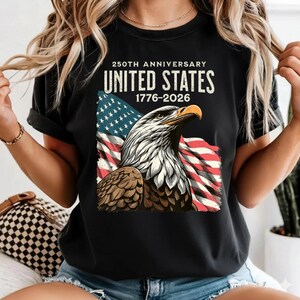 May include: Black t-shirt featuring a graphic of an American bald eagle with the US flag in the background. The text reads "250th Anniversary United States 1776-2026". The eagle is depicted in shades of brown, white, and gray.