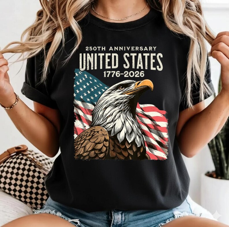 May include: Black t-shirt featuring a graphic of an American bald eagle with the US flag in the background. The text reads "250th Anniversary United States 1776-2026". The eagle is depicted in shades of brown, white, and gray.