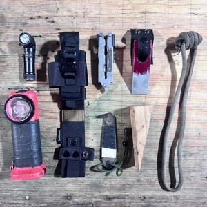 May include: A collection of outdoor gear including a black flashlight, a red and black headlamp, a black TacMount sheath, a silver multi-tool, a red and silver tool in a black holder, a small knife, a wooden wedge, and a long gray rope with a knot.