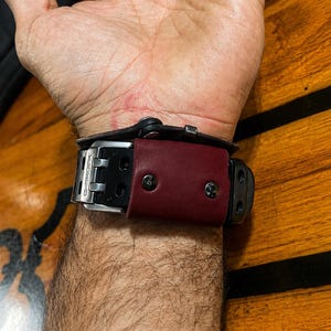 May include: A black watch with a metallic silver buckle and a dark red leather strap keeper secured by two black rivets. The watch band features the text G-SHOCK embossed on the silver metal buckle component, visible on the wrist.