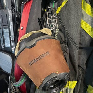 May include: A brown leather cover with the name MURCHISON embossed on the front, fitted over a black MSA respirator mask component. The item is attached to a black and yellow firefighter turnout coat with reflective strips and a red harness strap.