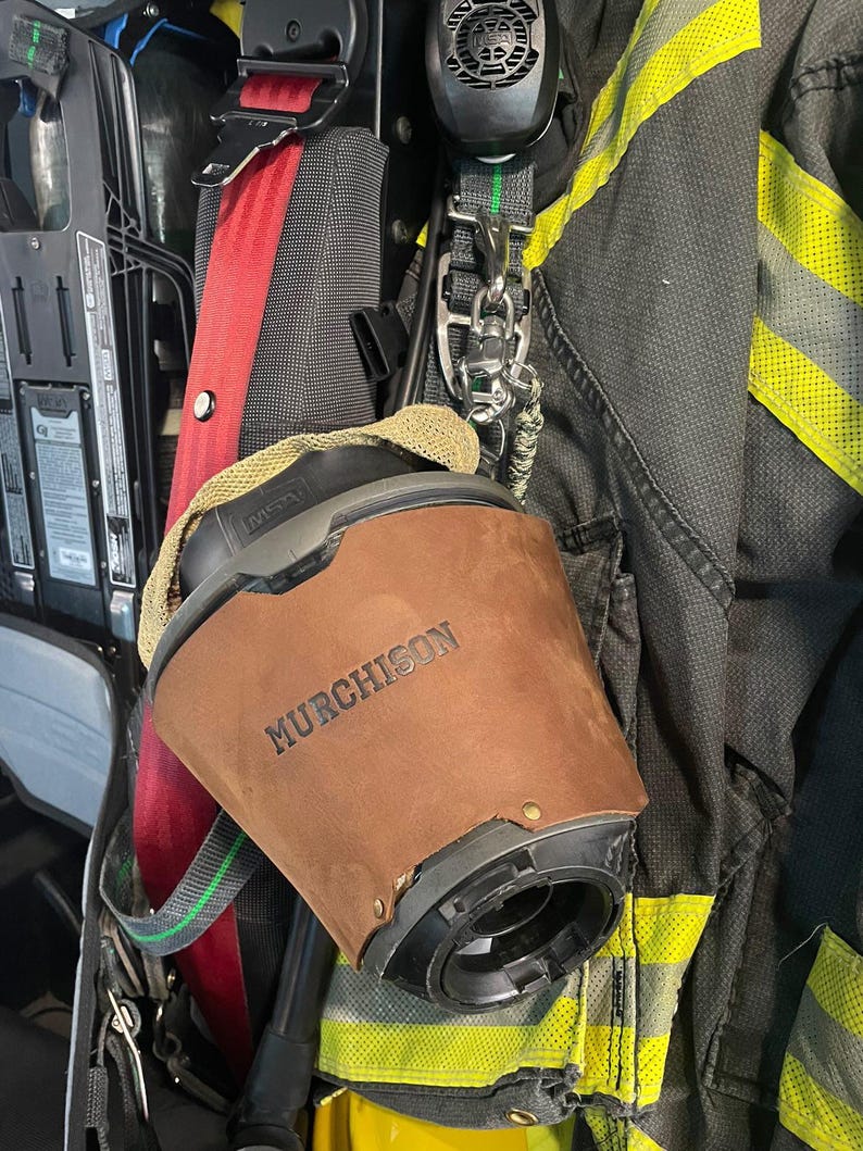 May include: A brown leather cover with the name MURCHISON embossed on the front, fitted over a black MSA respirator mask component. The item is attached to a black and yellow firefighter turnout coat with reflective strips and a red harness strap.
