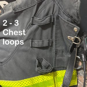 May include: A dark gray utility vest featuring multiple fabric chest loops, a silver metal D-ring, and a bright yellow reflective strip along the bottom edge. The text 2 - 3 Chest loops is printed in white on the left side of the garment.