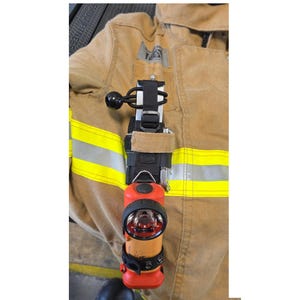 May include: A tan firefighter jacket with horizontal reflective yellow stripes features a black nylon strap mount attached to the chest. The mount holds a bright orange flashlight with a black circular lens and a black elastic cord fastener.