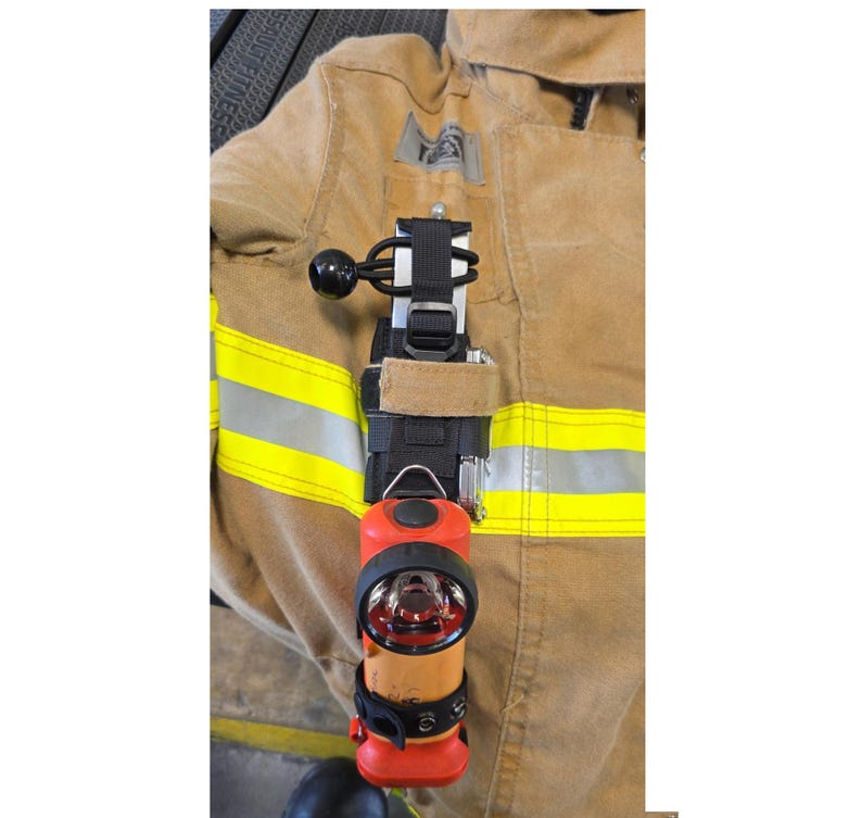 May include: A tan firefighter jacket with horizontal reflective yellow stripes features a black nylon strap mount attached to the chest. The mount holds a bright orange flashlight with a black circular lens and a black elastic cord fastener.