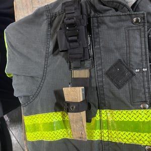 May include: A dark gray firefighter turnout jacket featuring a horizontal neon yellow reflective strip, a black nylon gear strap, and a wooden door wedge secured in a black fabric holster attached to the front of the garment.