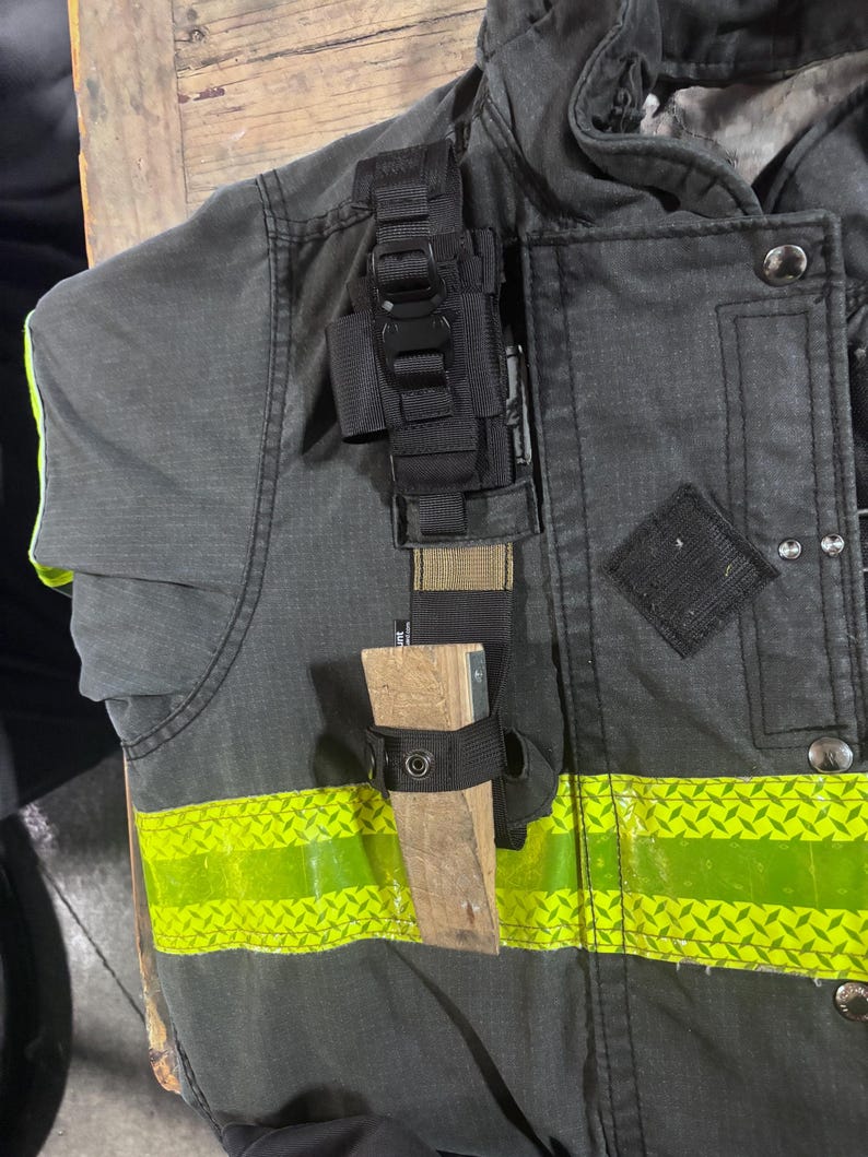 May include: A dark gray firefighter turnout jacket featuring a horizontal neon yellow reflective strip, a black nylon gear strap, and a wooden door wedge secured in a black fabric holster attached to the front of the garment.