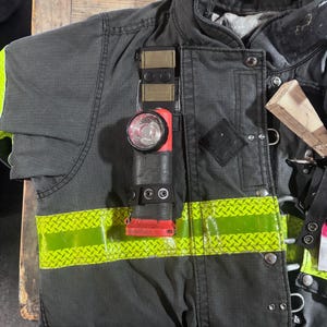 May include: A dark gray firefighter turnout coat featuring a horizontal lime green reflective strip with a diamond pattern. A red and black flashlight is attached to the chest area with a tan and black strap, secured by two black metal snaps.