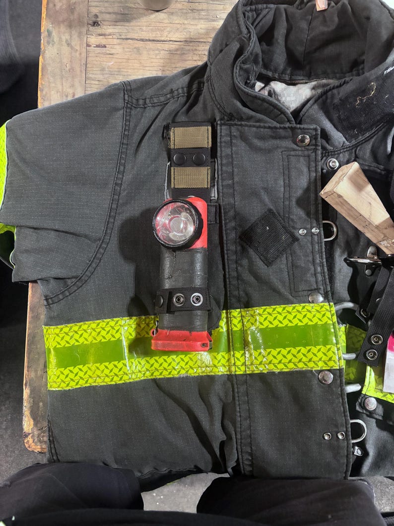 May include: A dark gray firefighter turnout coat featuring a horizontal lime green reflective strip with a diamond pattern. A red and black flashlight is attached to the chest area with a tan and black strap, secured by two black metal snaps.