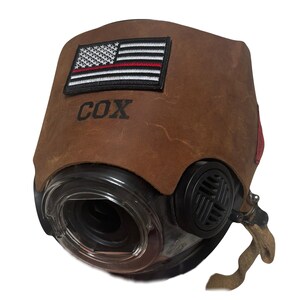 May include: A brown leather cover for a respirator mask featuring an embroidered American flag patch with a thin red line. The name COX is embossed in black lettering below the flag. The mask has a clear circular lens and black side vents.