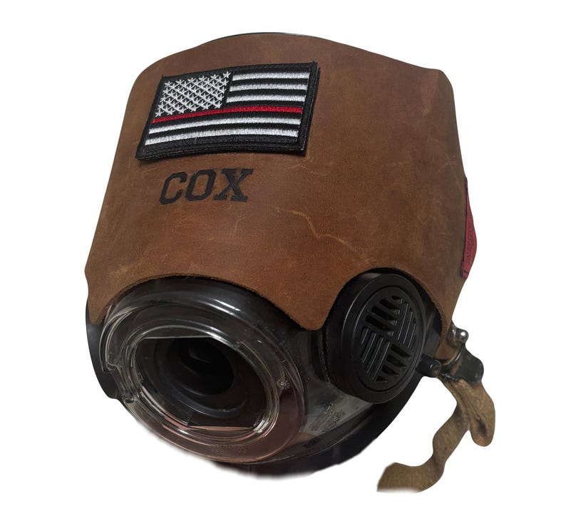 May include: A brown leather cover for a respirator mask featuring an embroidered American flag patch with a thin red line. The name COX is embossed in black lettering below the flag. The mask has a clear circular lens and black side vents.
