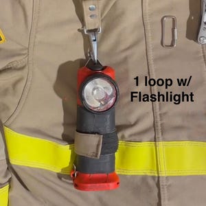 May include: A red and black flashlight attached to a tan fabric loop with a metal clip, secured to a tan jacket with yellow reflective strips. The text 1 loop w/ Flashlight is printed next to the flashlight on the tan fabric background.