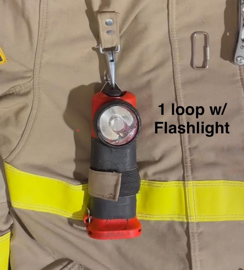 May include: A red and black flashlight attached to a tan fabric loop with a metal clip, secured to a tan jacket with yellow reflective strips. The text 1 loop w/ Flashlight is printed next to the flashlight on the tan fabric background.