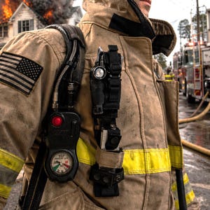 May include: A tan firefighter turnout coat with yellow reflective stripes and a black American flag patch on the shoulder. The coat features a black pressure gauge with a red button and a black utility pouch holding a flashlight and tools.