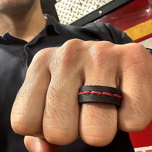 May include: A black leather ring with red stitching. The ring is worn on a finger and has a thick band. The stitching is visible across the top of the ring, adding a contrasting color element to the design. The ring appears to be handmade.