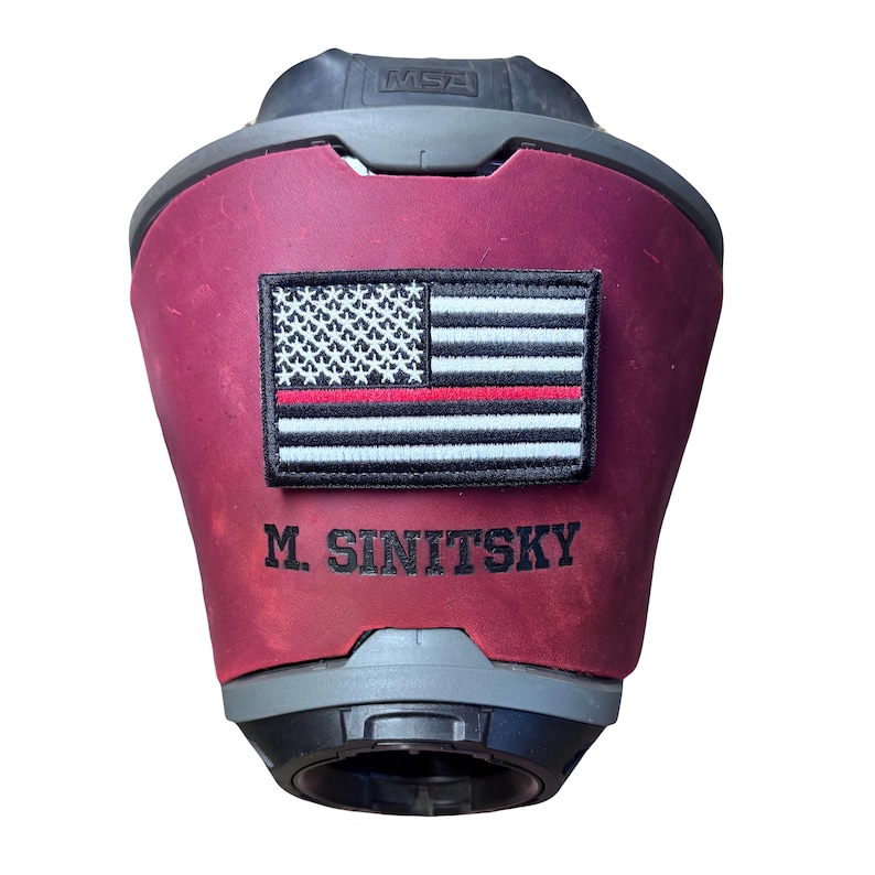 May include: A maroon leather cover for an MSA helmet featuring a rectangular patch with a black and white American flag design and a thin red horizontal line. The name M. SINITSKY is embroidered in black thread below the flag patch.