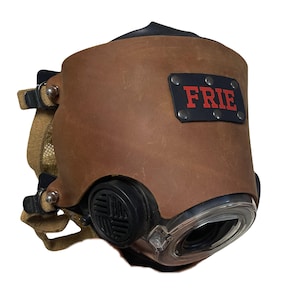 May include: A brown leather face mask featuring a black patch with the red text "FRIE" attached by four silver rivets. The mask includes a clear circular lens, a black circular side filter, and tan fabric straps with metal adjustment buckles.